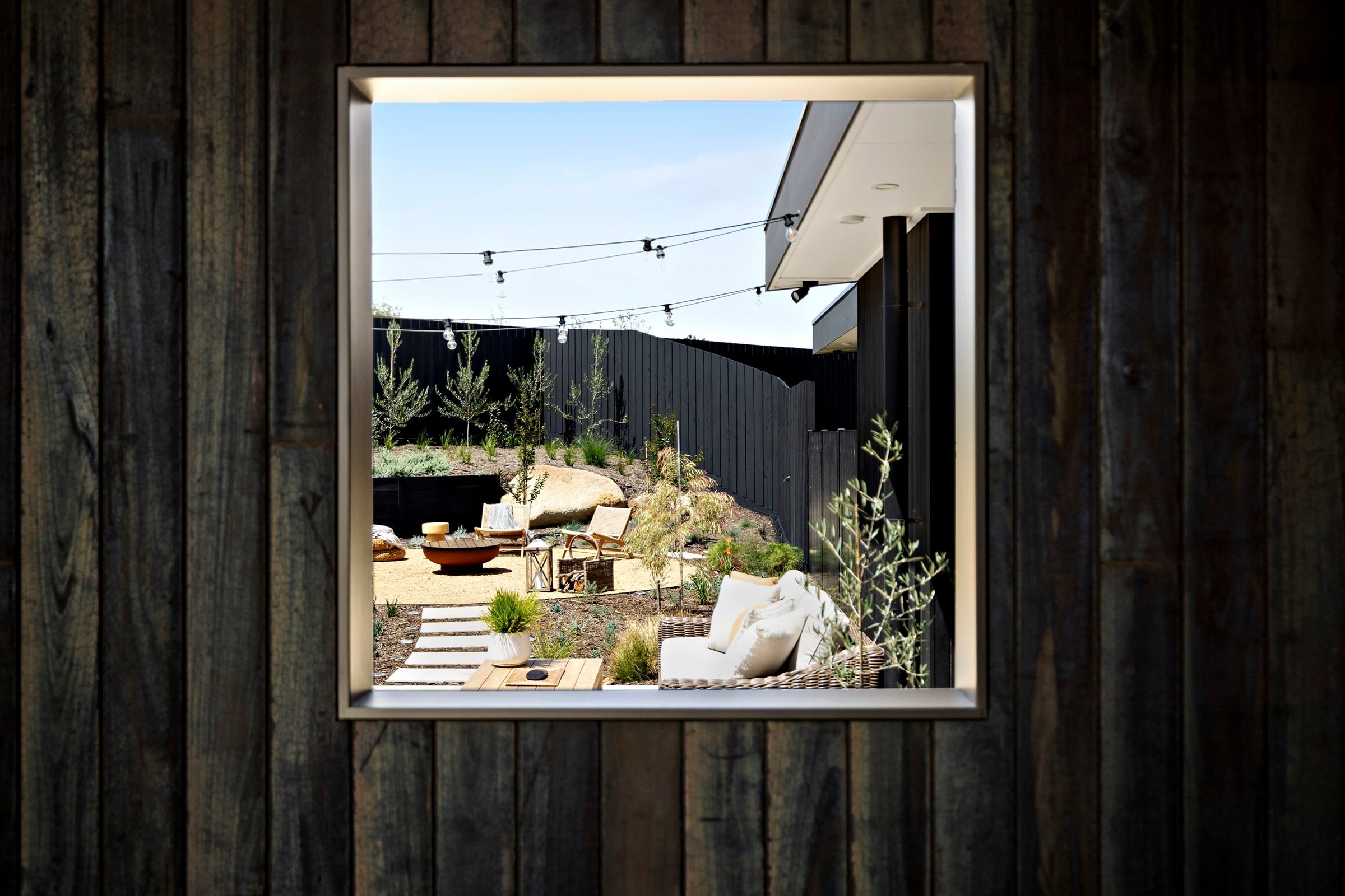 A dark-clad house with a large picture window looking out over an outdoor firepit at dusk.
