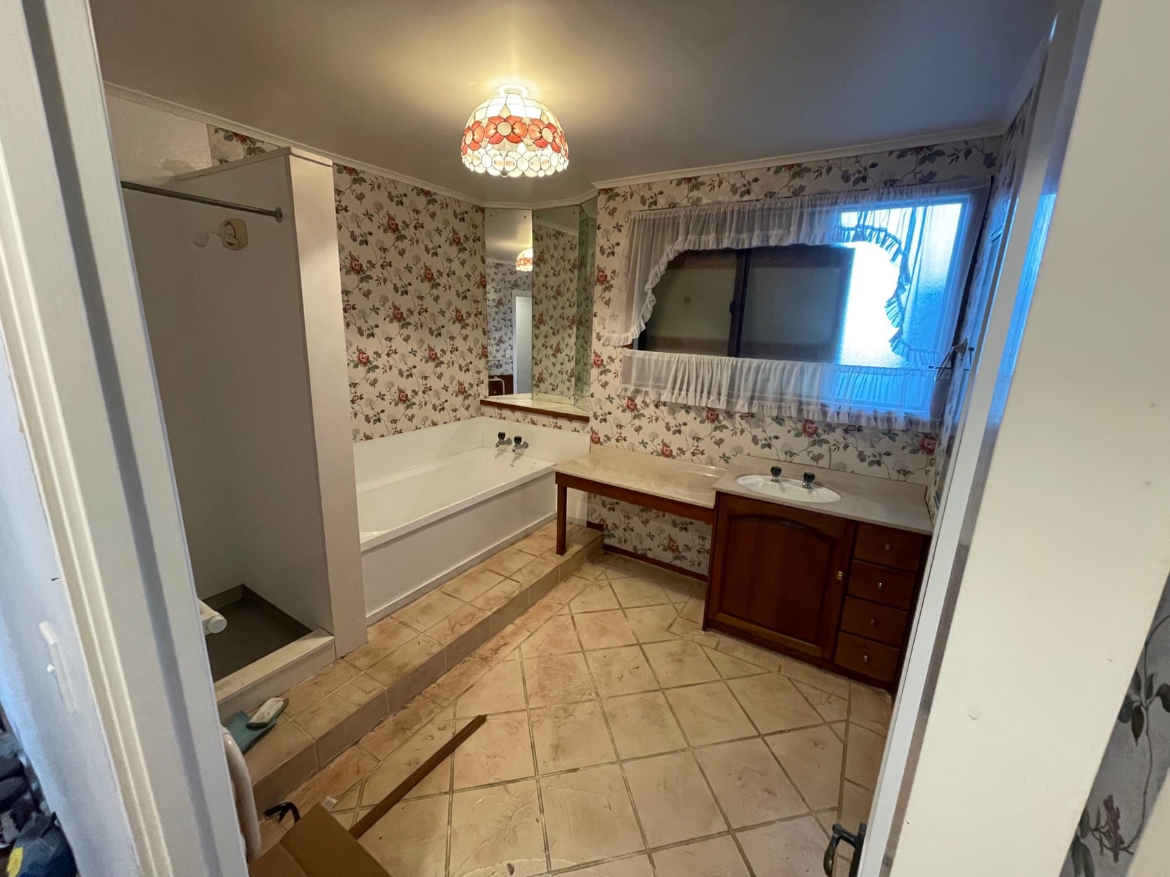 A dated bathroom before renovation, with patterned wallpaper, tiled floor and the original vanity.