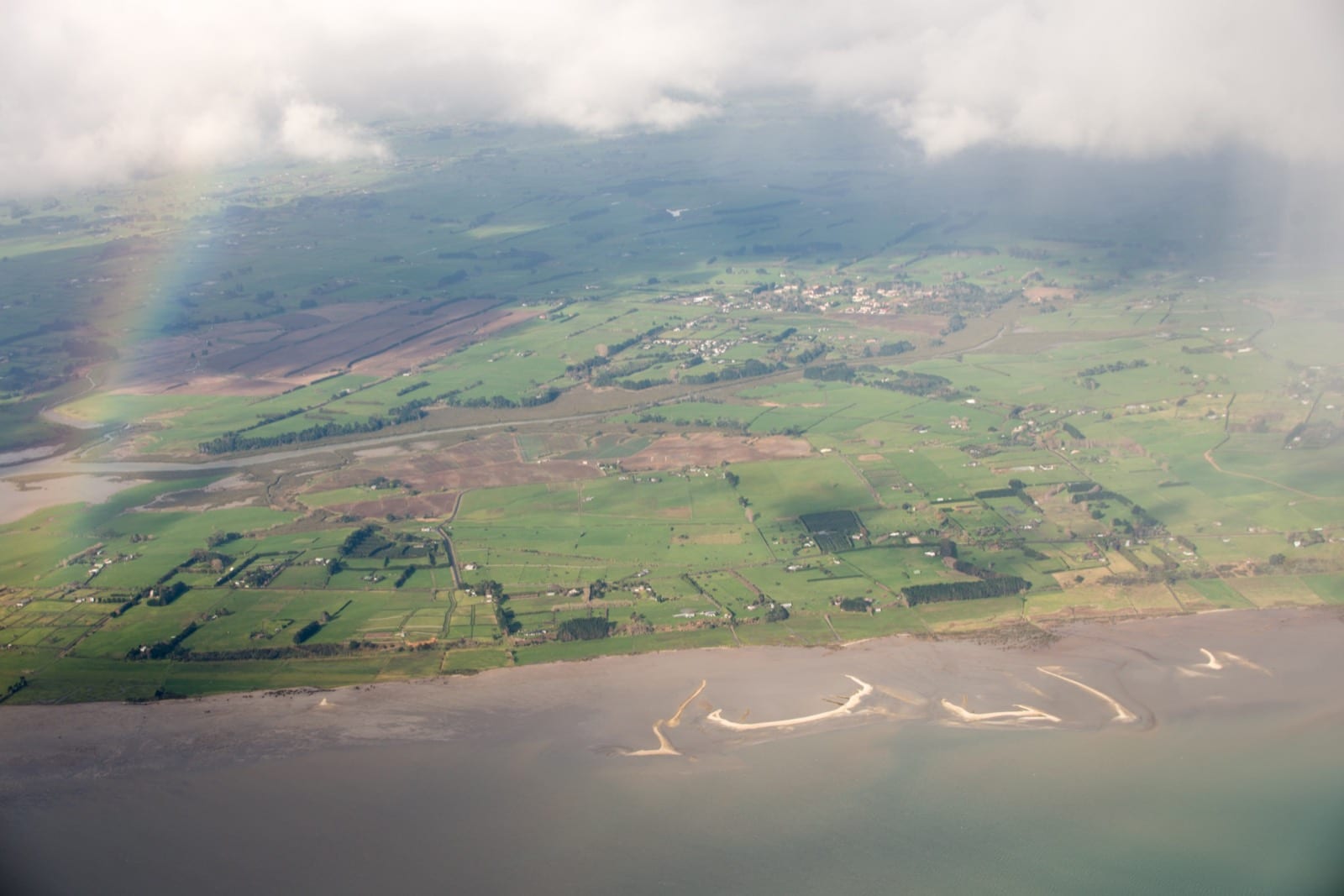 Waiau Pa farmland, the settlement and the Manukau Harbour shoreline with sandbars and a rainbow, from the air.
